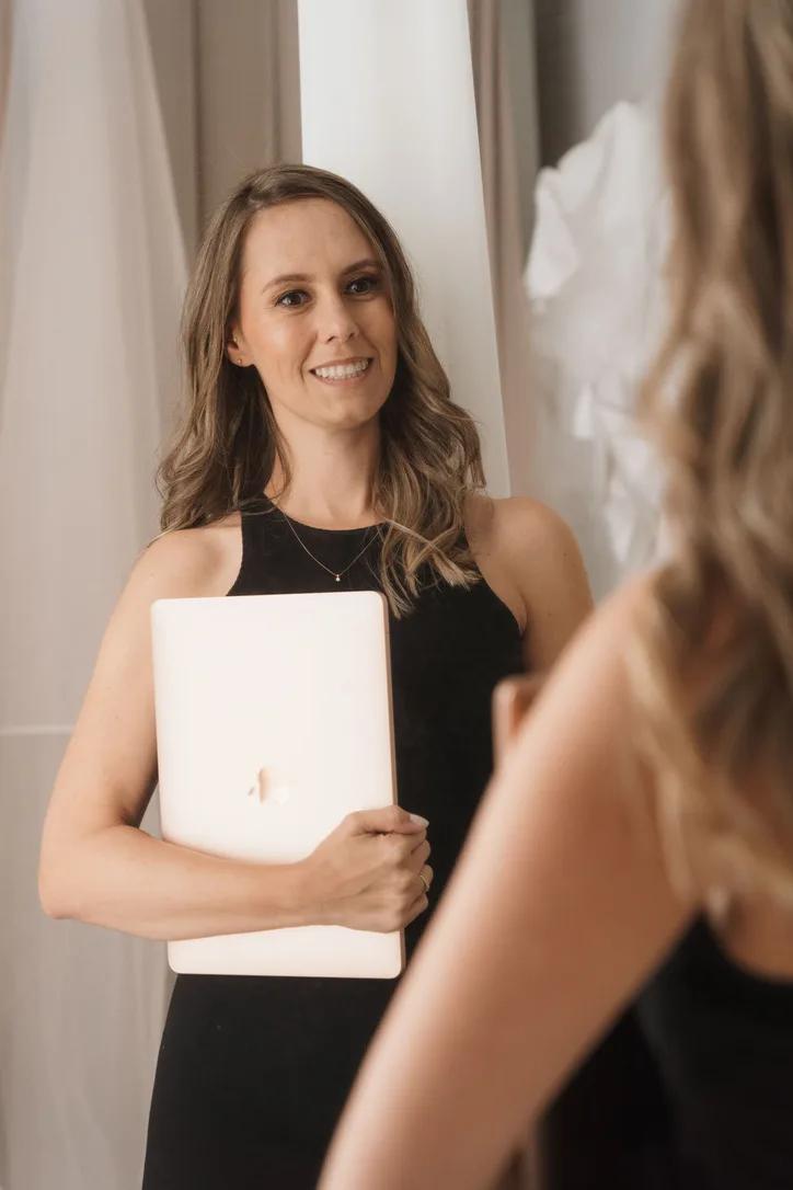 Jackie in a black dress, holding her laptop and smiling at her reflection